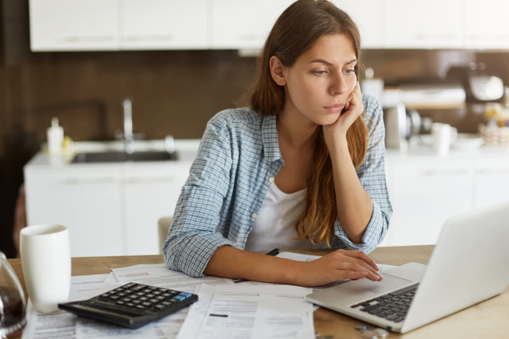 people, accounting, finances, family budget and financial issues concept. serious young european woman calculating domestic expenses, sitting at dining table in front of open laptop computer